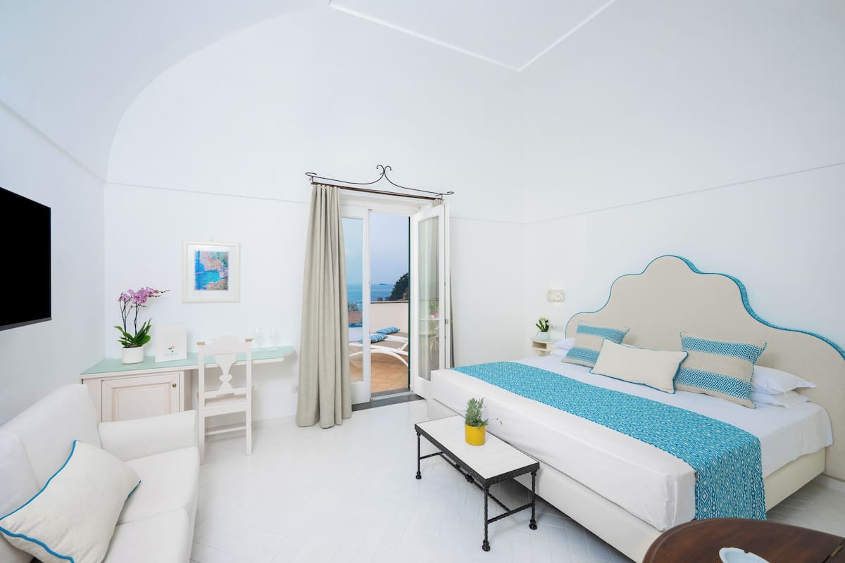 A bedroom with a bed, a desk, a chair, a small table, and a view of the outdoors.