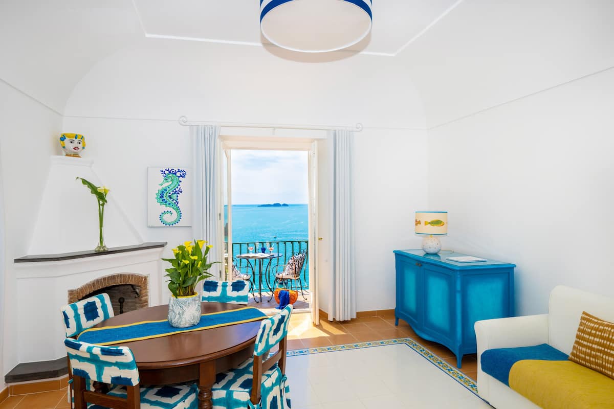 A room with a dining table, chairs, a sofa, and a view of the sea.