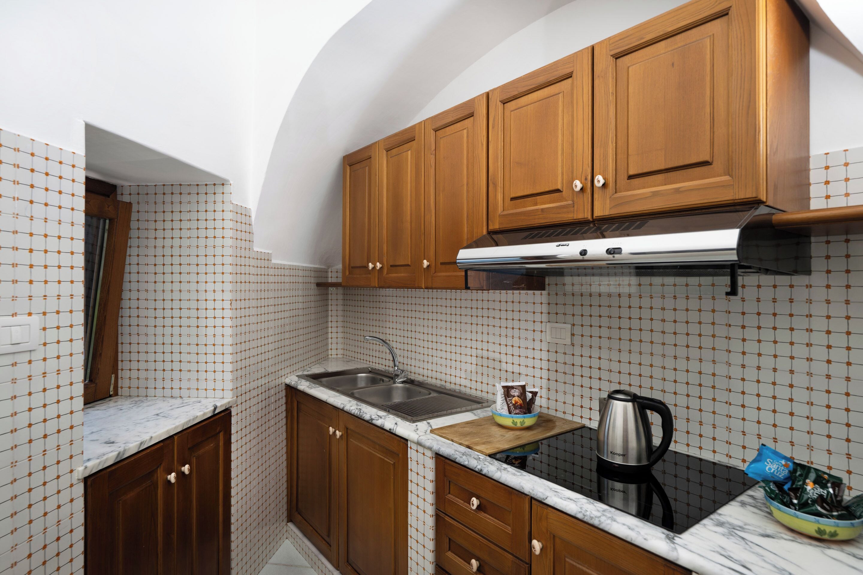 A kitchen with wooden cabinets, a stainless steel sink, a marble countertop, and a kettle.