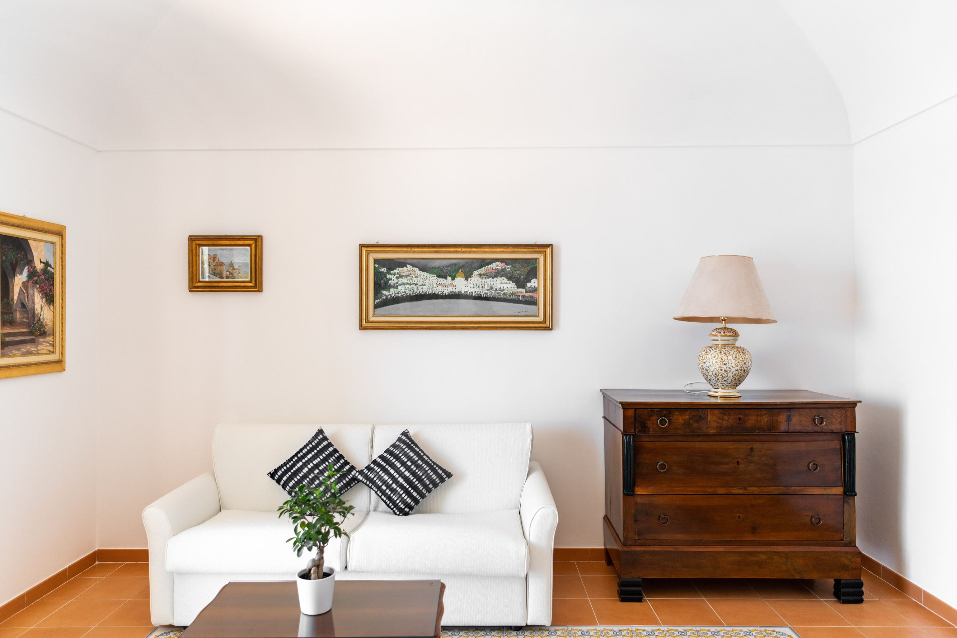 A white sofa with black and white patterned pillows, a wooden cabinet with a lamp, and two framed pictures on the wall.