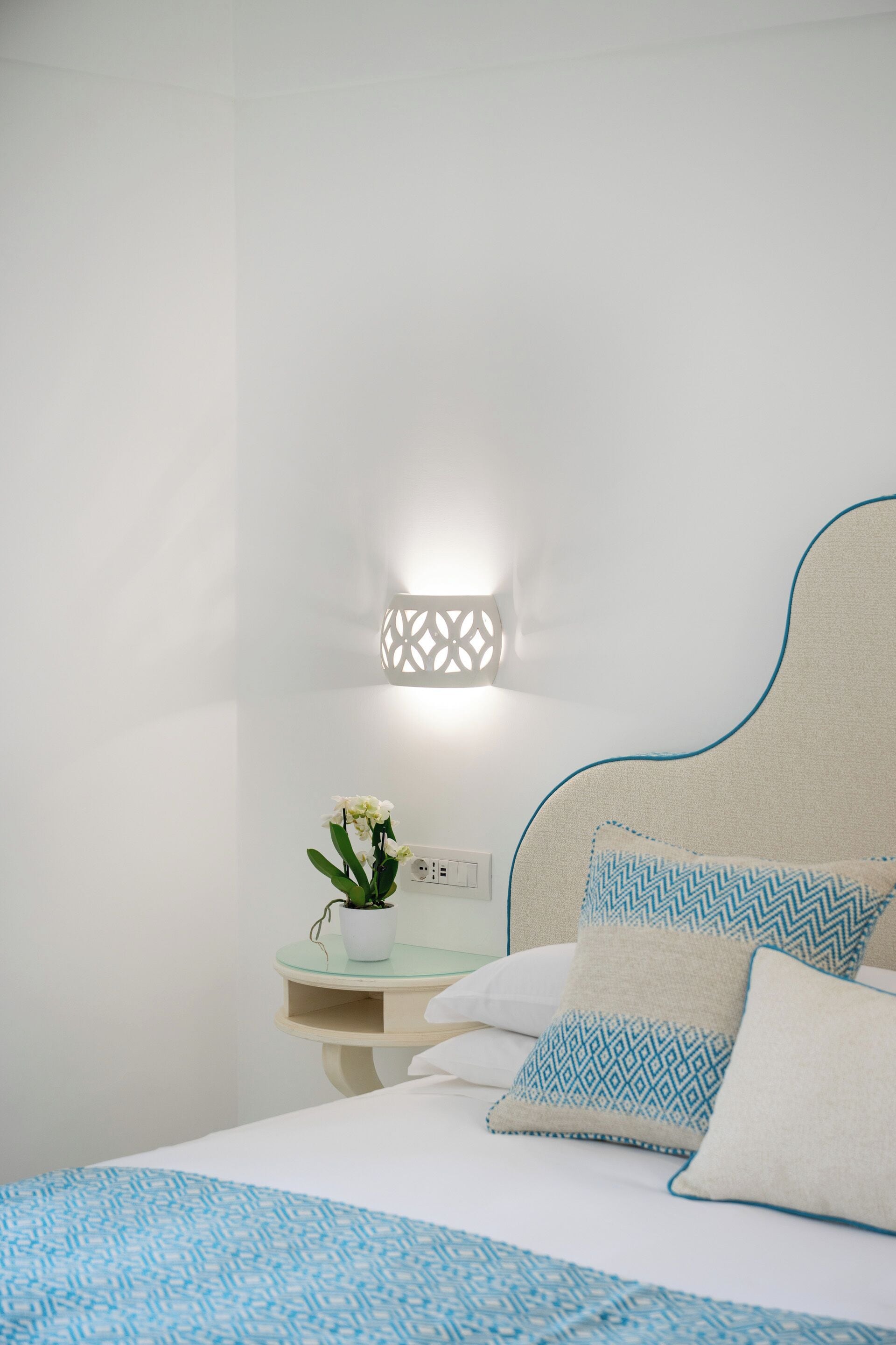 A neatly made bed with patterned pillows, a bedside table with a potted plant, and a wall-mounted light fixture.