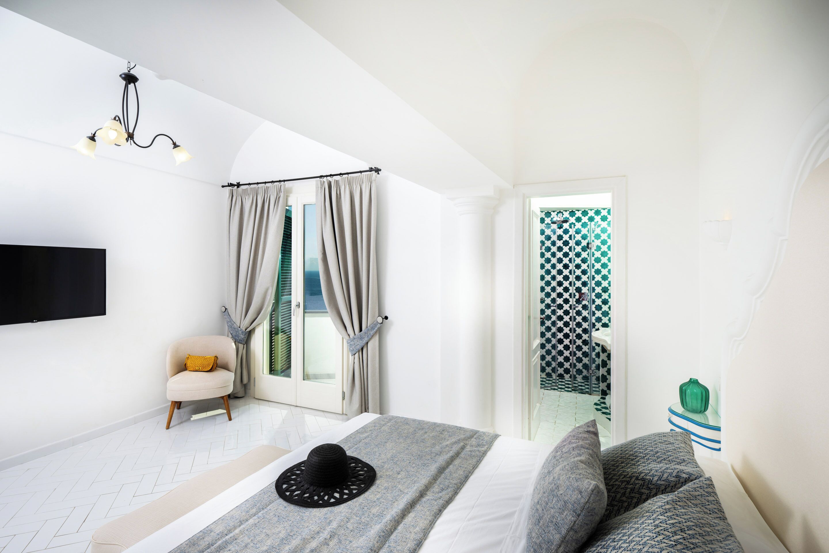 A bedroom with a bed, a bench, a chair, a TV, a chandelier, and a view of a balcony.