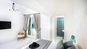 A bedroom with a bed, a bench, a chair, a TV, a chandelier, and a view of a balcony.