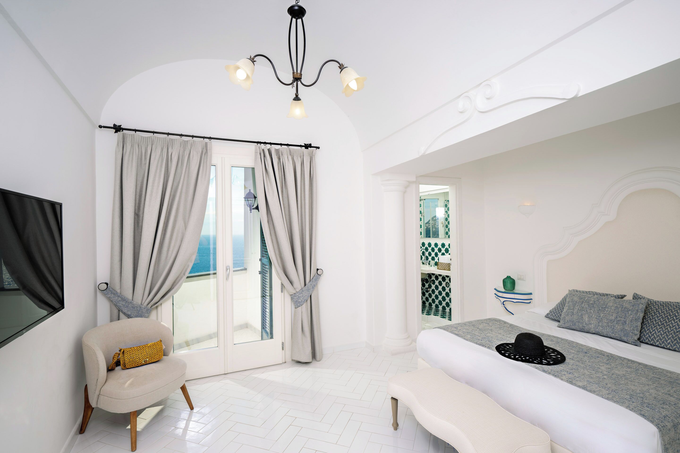 Een slaapkamer met een bed, een bankje, een stoel, een kroonluchter en uitzicht op zee.