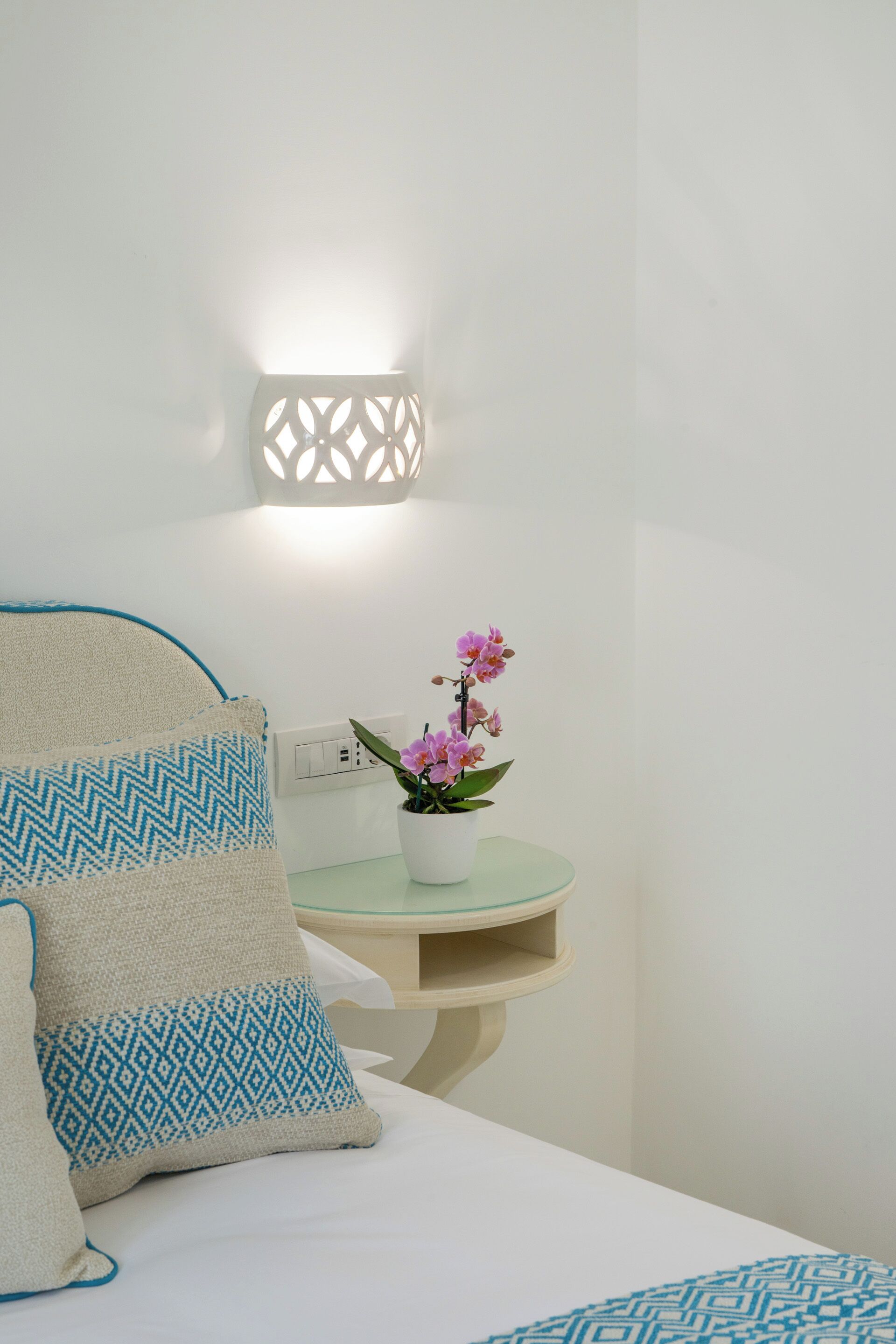 A neatly arranged bedroom with a bed, pillows, a bedside table with a potted orchid, and a wall-mounted light fixture.