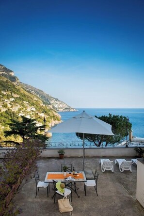 A patio with a table and chairs, a white umbrella, and a view of the sea and hills. - Alcione Residence (Positano)