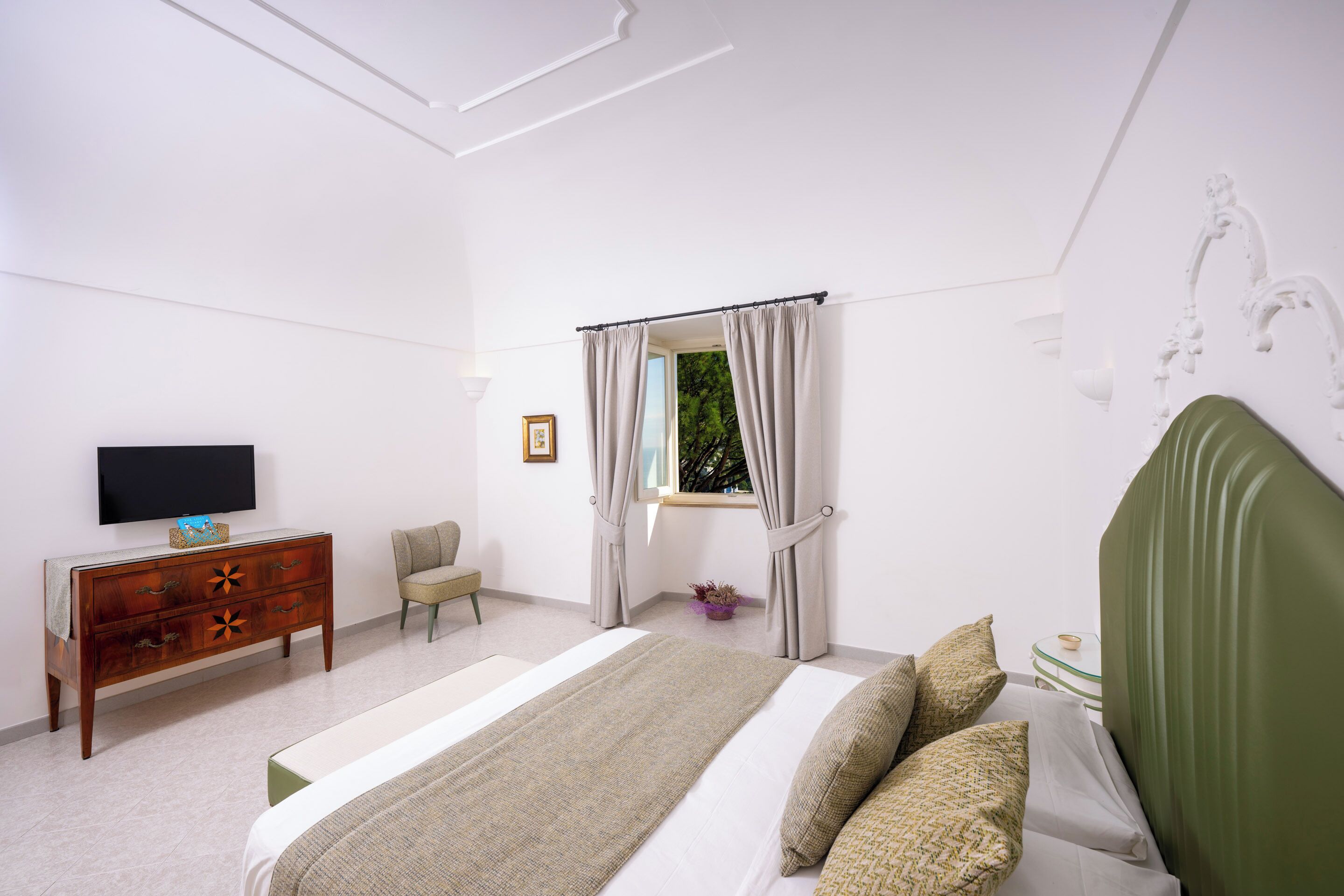 Une chambre à coucher avec un lit, une commode, un téléviseur, une fenêtre avec des rideaux et une plante.