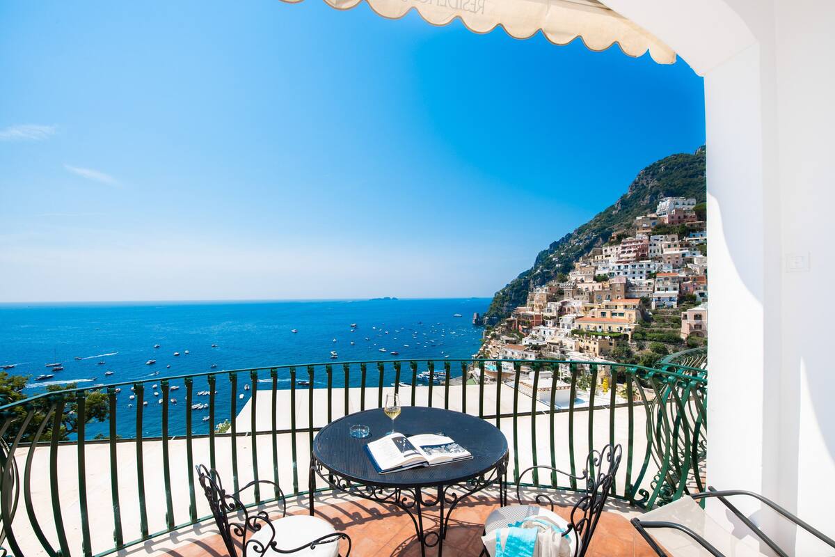 A balcony with a table and chairs overlooking a coastal town and the sea.