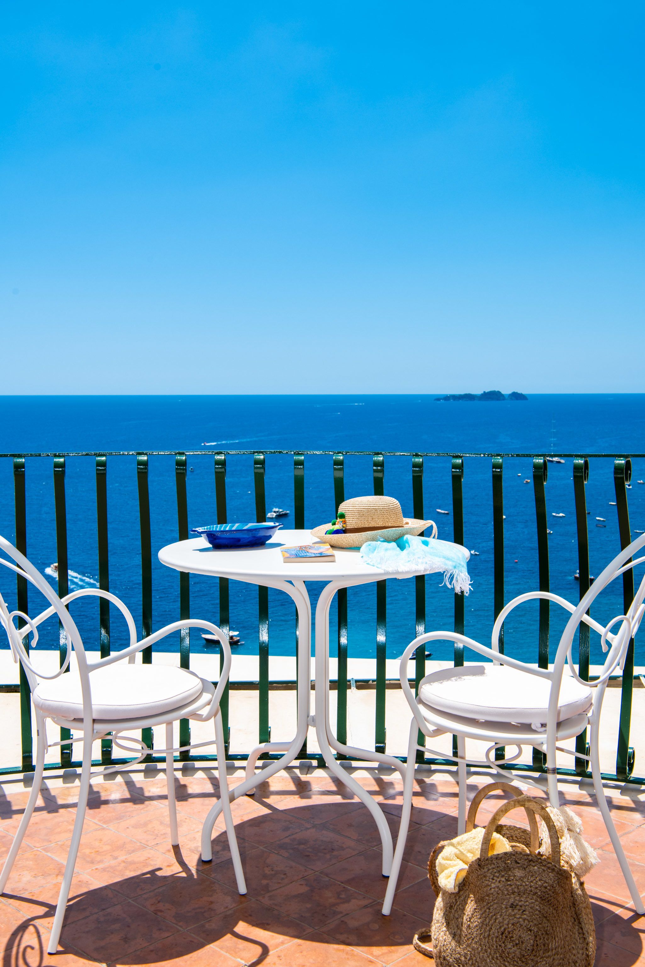 A terrace with two white chairs and a table, overlooking a clear blue sea with boats.