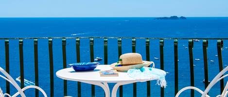A terrace with two white chairs and a table, overlooking a clear blue sea with boats.
