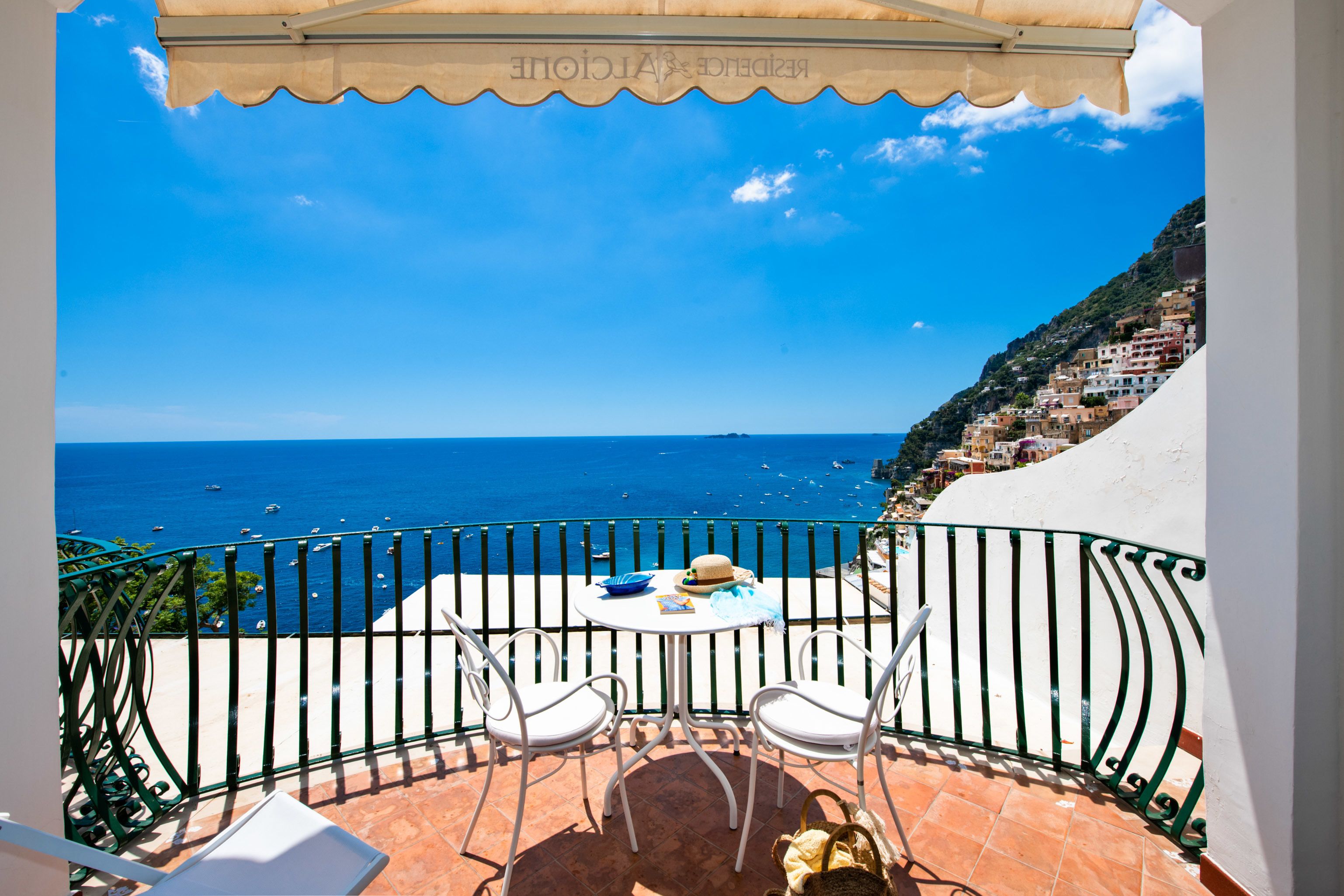 A balcony with a view of the sea, white chairs, and a table with a hat and book.