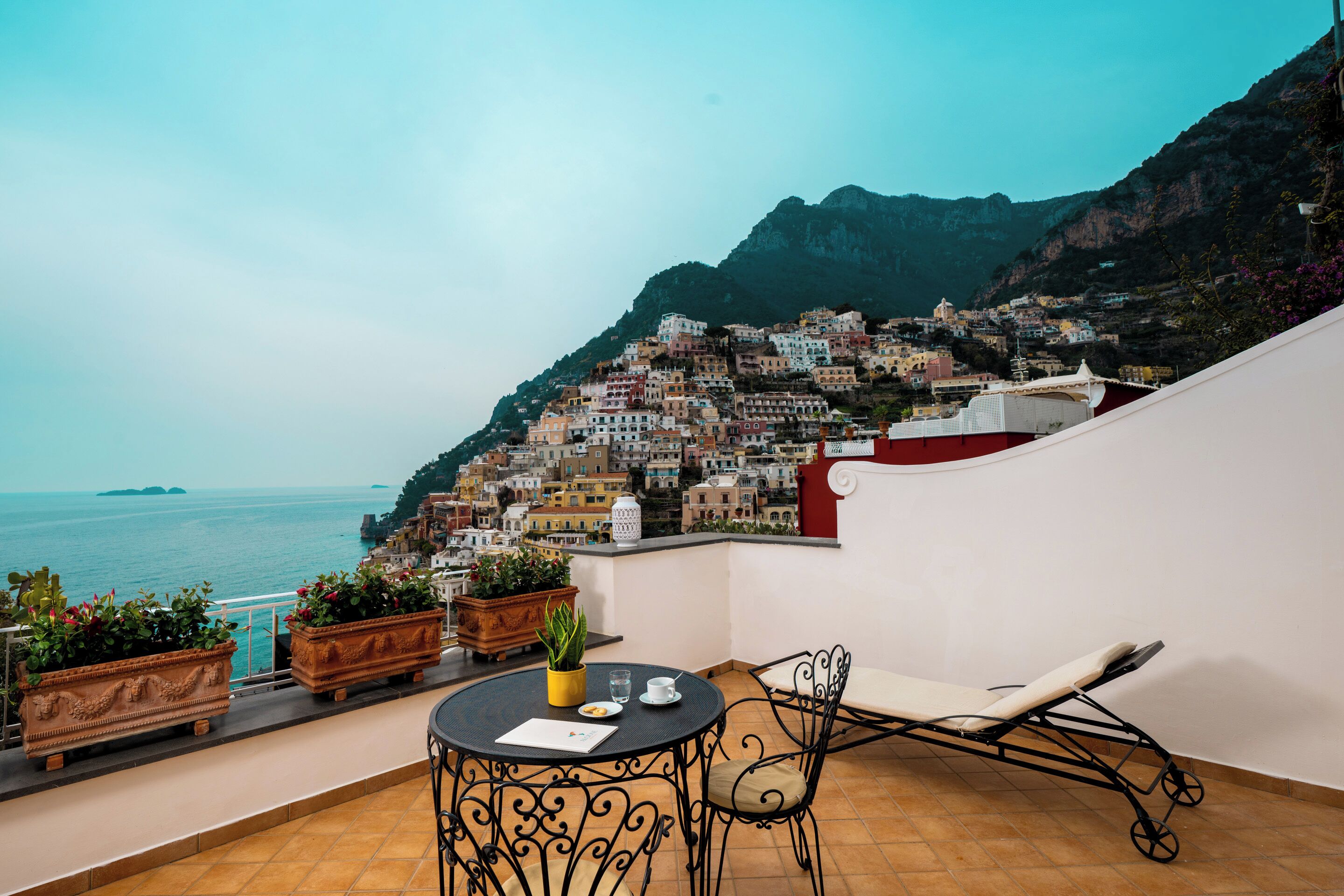 A terrace with a table, chairs, potted plants, and a view of a coastal town.
