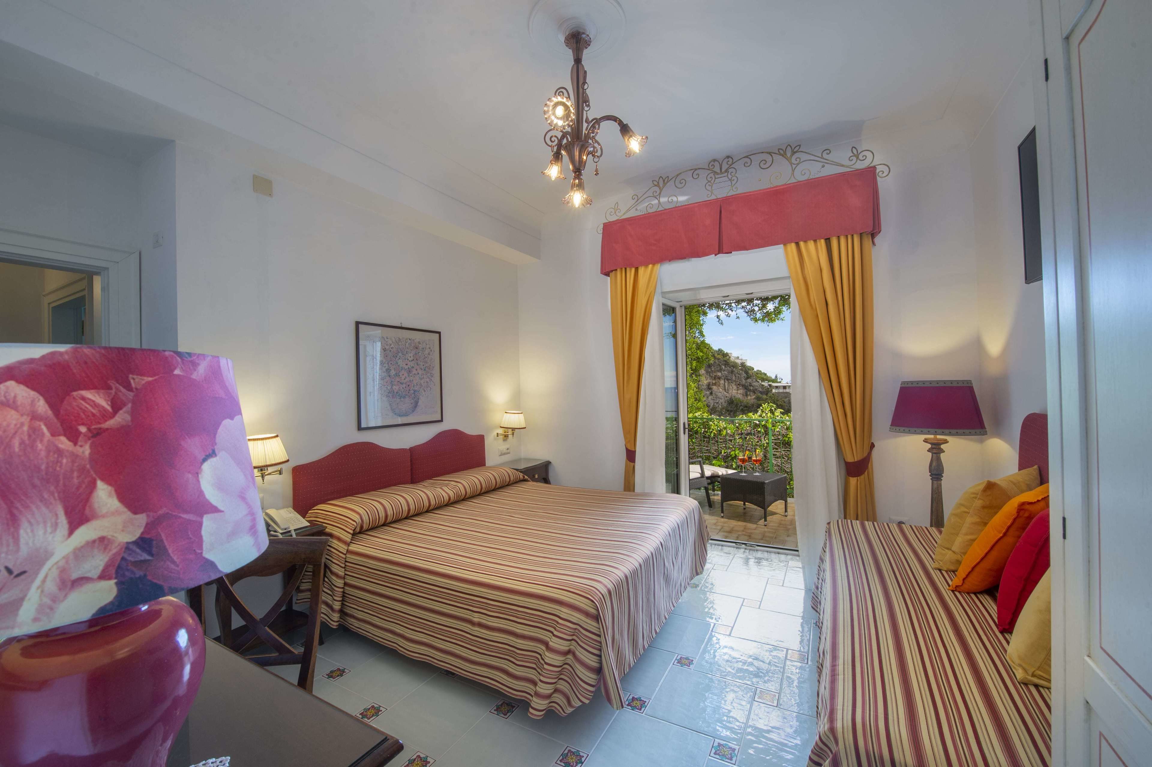 Une chambre d’hôtel avec un lit, un bureau et une chaise. Il y a une fenêtre avec des rideaux, un lustre et une vue sur l’extérieur.