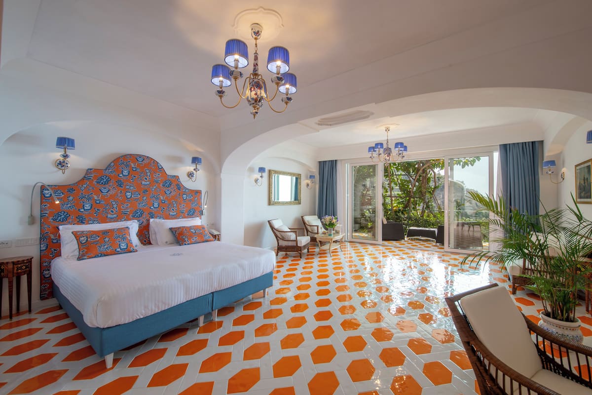 A hotel room with a large bed, a chandelier, a balcony with a view, and a patterned floor.