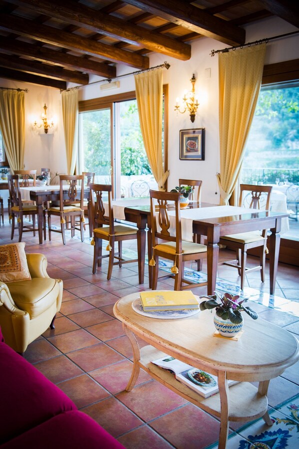 A dining area with wooden tables and chairs, a yellow sofa, and a potted plant on a table. - Hotel Pellegrino (Praiano)