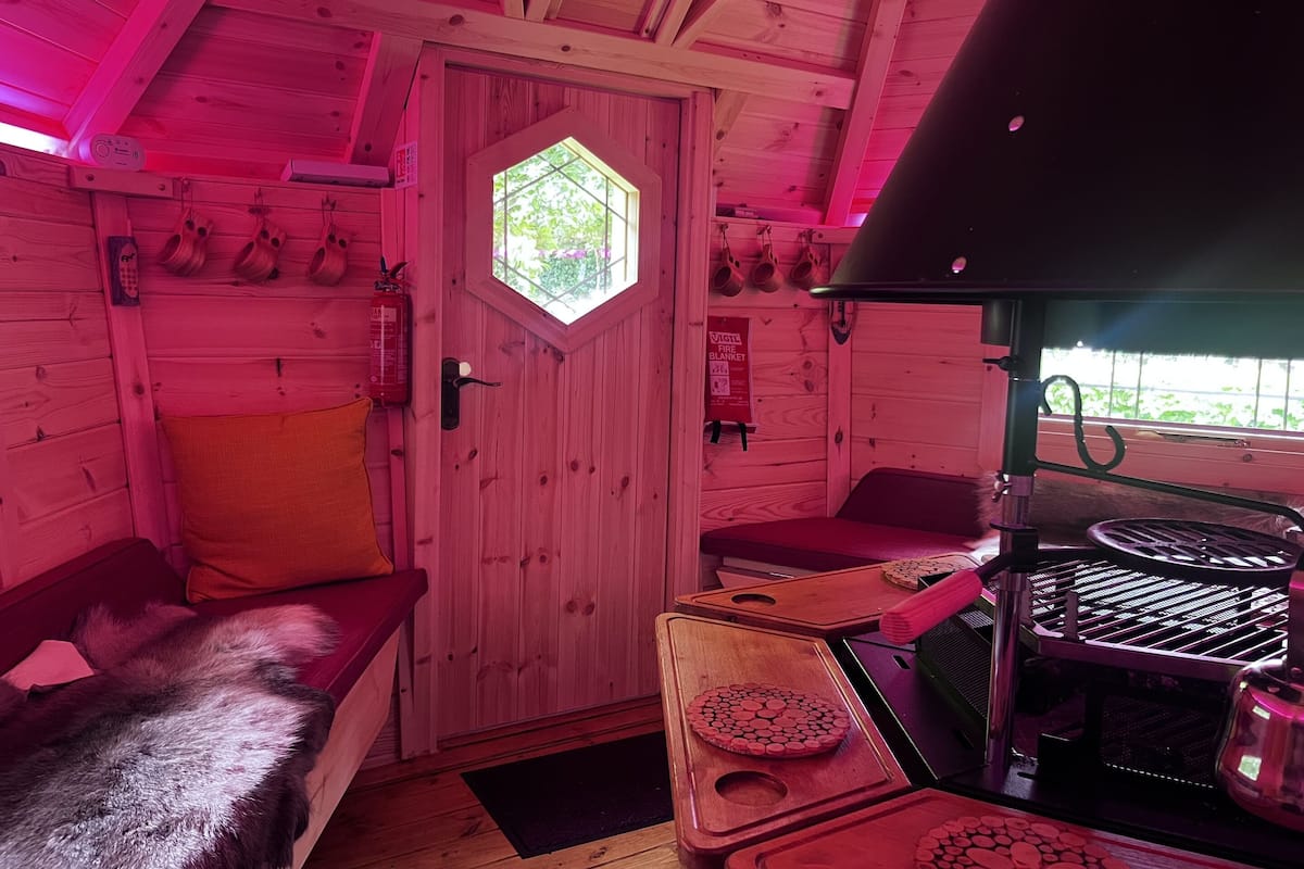 Interior de una cabaña con una puerta de madera, un banco, una parrilla y una ventana.