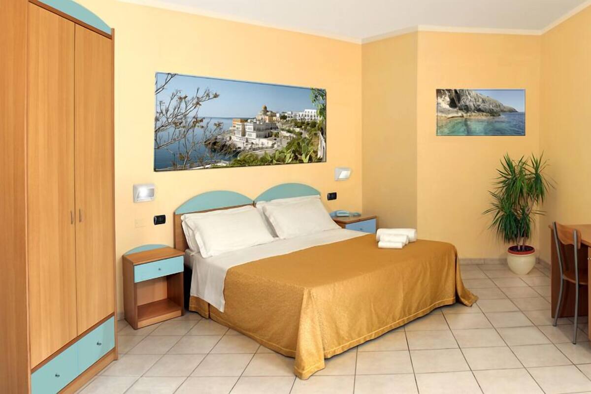 A hotel room with a bed, a desk with a chair, a TV, and two paintings on the wall.