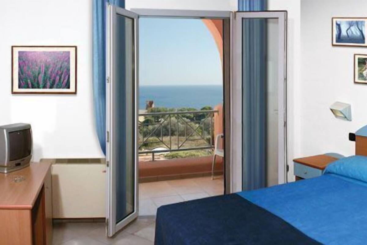 A hotel room with a bed, a desk with a television, a balcony with a view of the sea, and framed pictures on the wall.