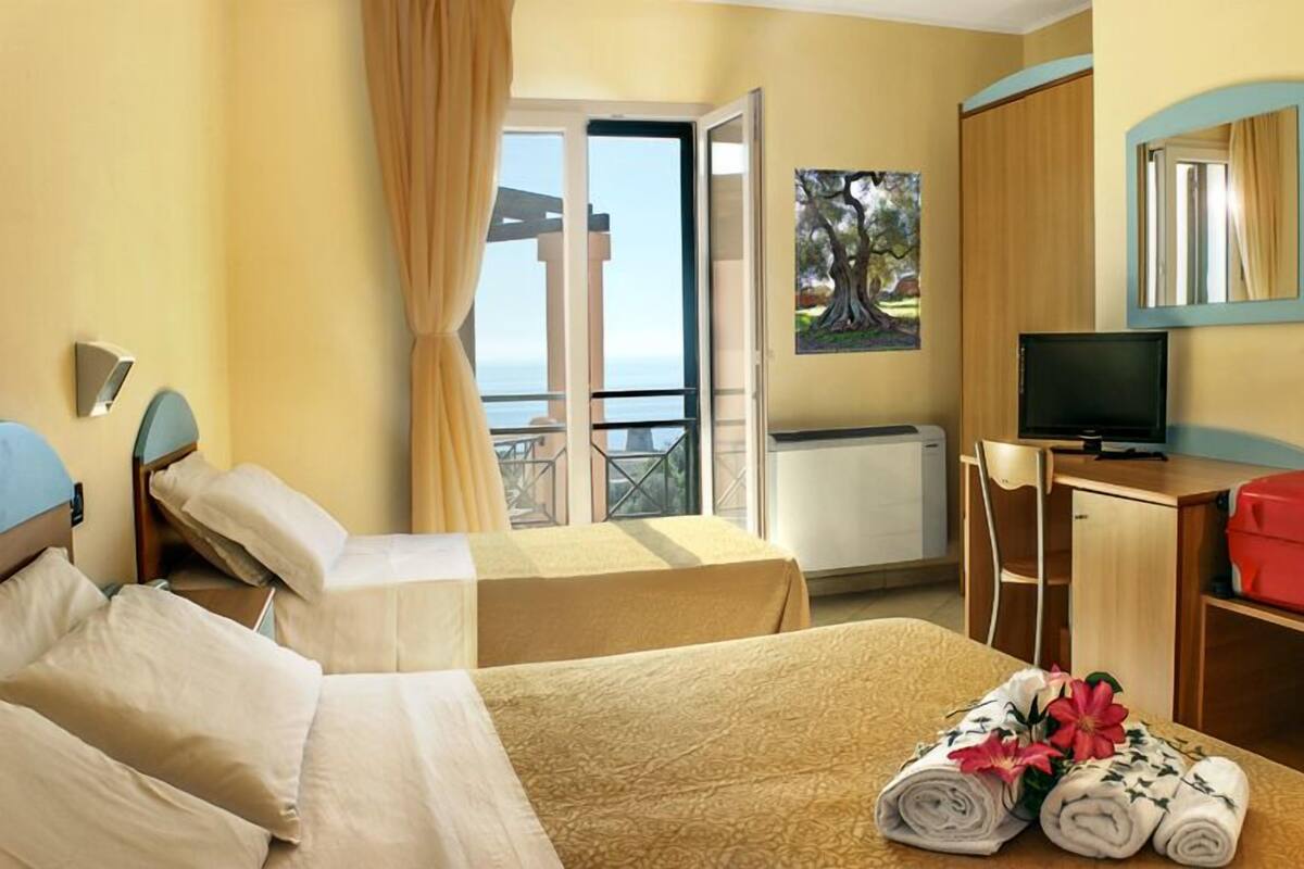 A hotel room with two beds, a desk with a TV, a red suitcase, and a balcony with a view.