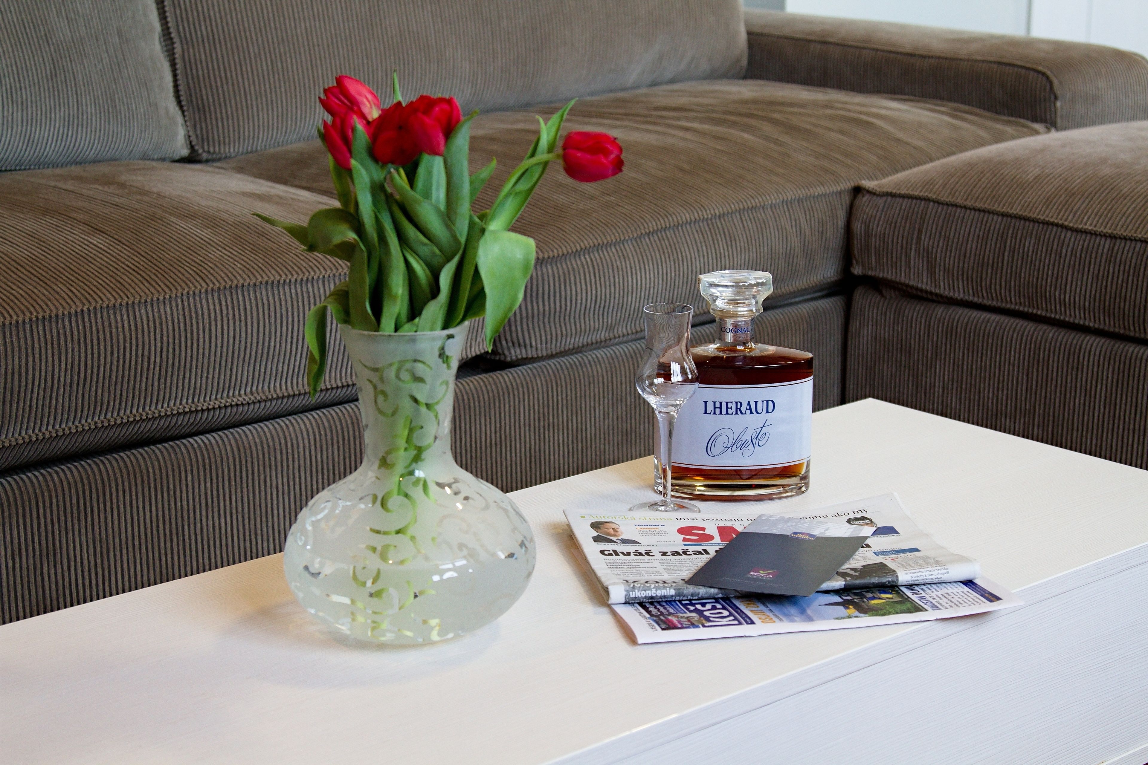 Uma mesa branca com um vaso de vidro contendo tulipas vermelhas, uma garrafa de L'HERAU e um jornal.