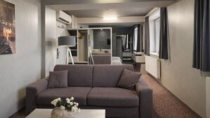 A modern hotel room with a grey sofa, a small table with a vase, a floor lamp, and a view of the hallway.