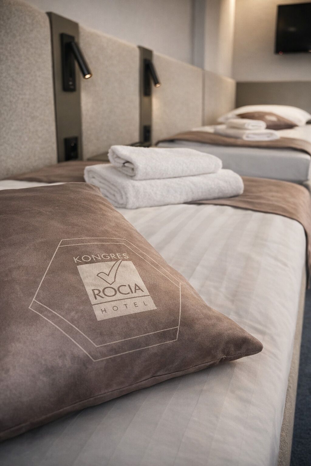 Habitación de hotel con una cama que incluye una almohada con el logotipo del hotel, cabecero con iluminación integrada y toallas dobladas con cuidado.