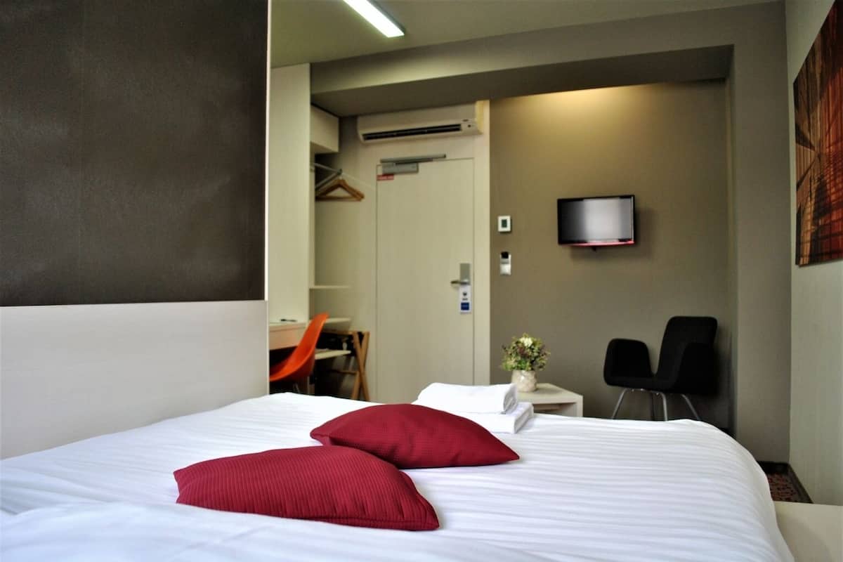 Hotellihuone, jossa on suuri sänky, kaksi punaista tyynyä, työpöytä tuolilla, seinälle asennettu televisio ja pieni pöytä, jossa on maljakko täynnä kukkia.