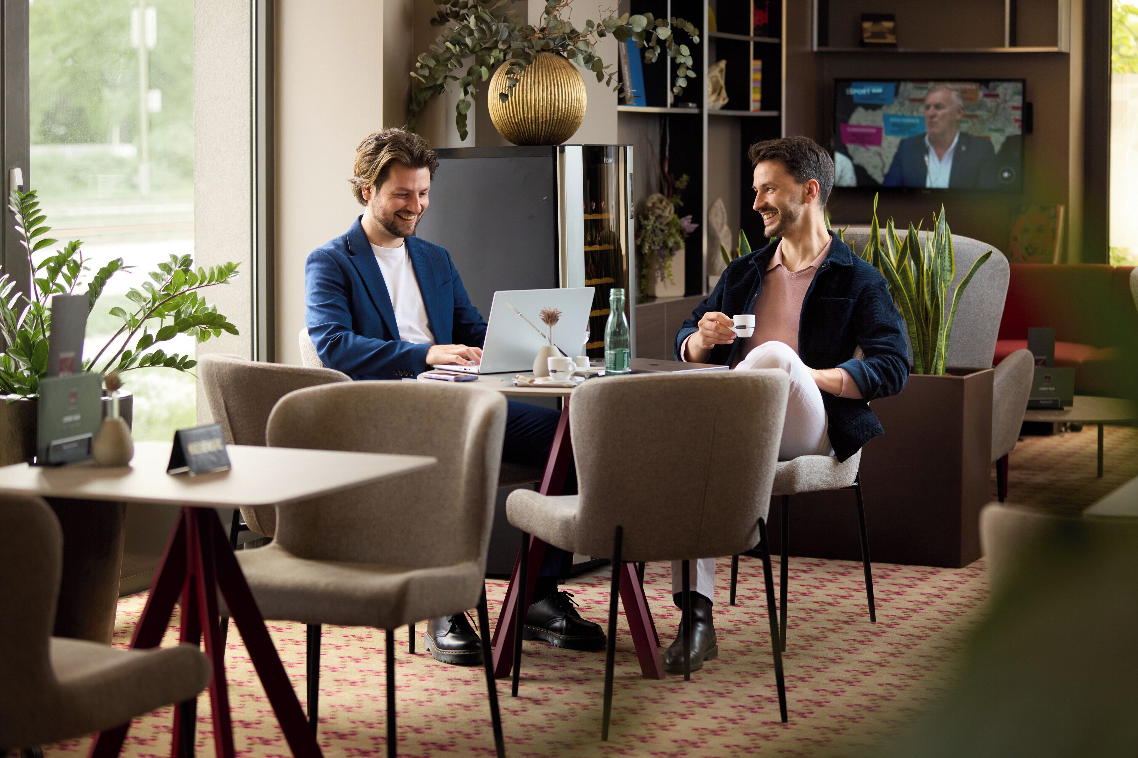 Dos hombres sentados a una mesa con computadoras portátiles, una taza de café y un florero con flores secas.