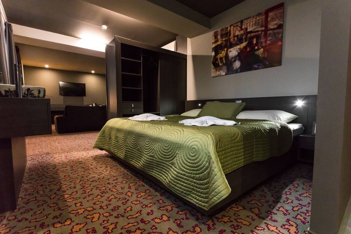 Moderni hotellihuone, jossa on suuri sänky, taulutelevisio, erillinen vaatehuone ja kaupunkimaisemaa esittävä taulu seinällä.