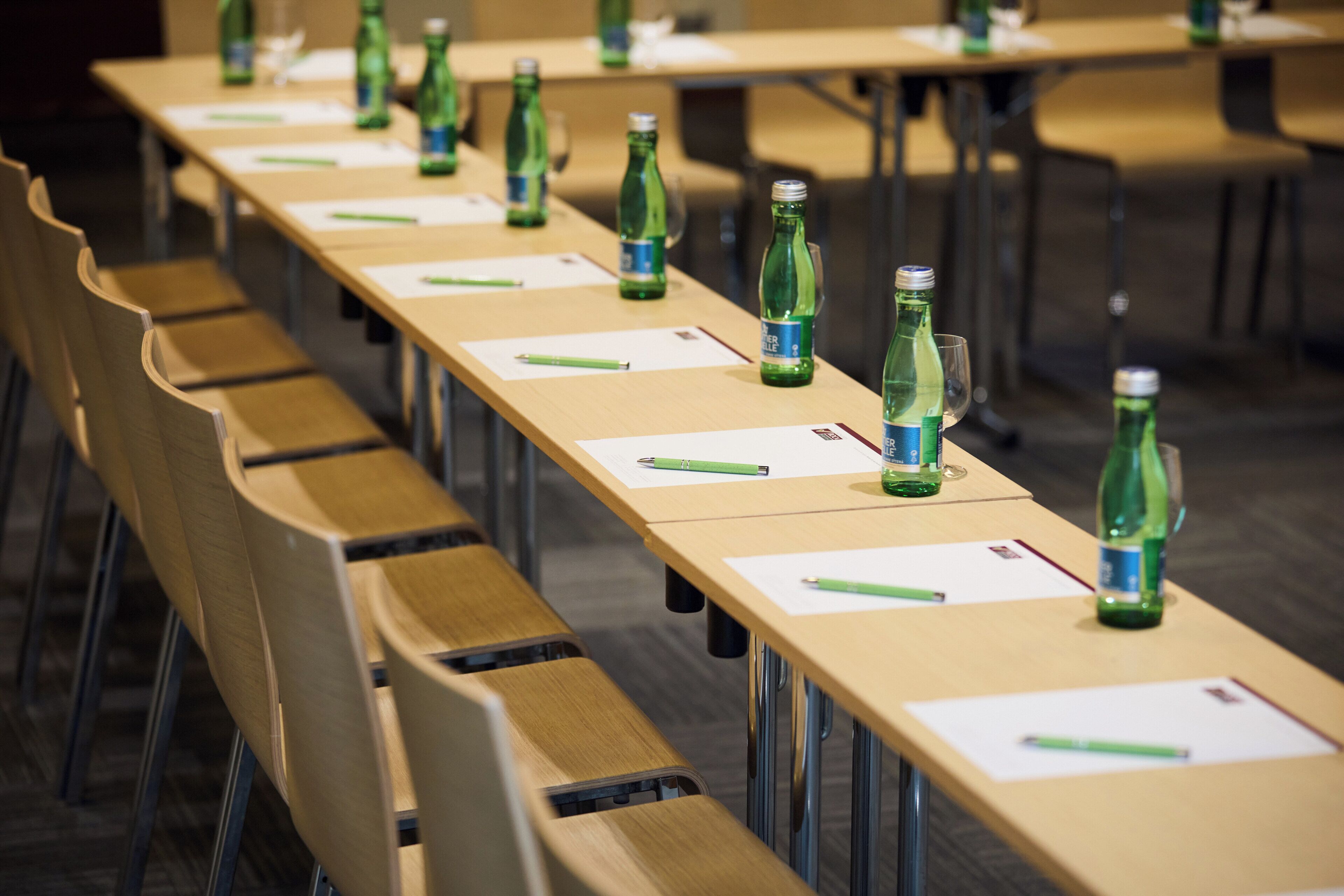 Un salón de conferencias con largas mesas, sillas y botellas verdes en cada una.