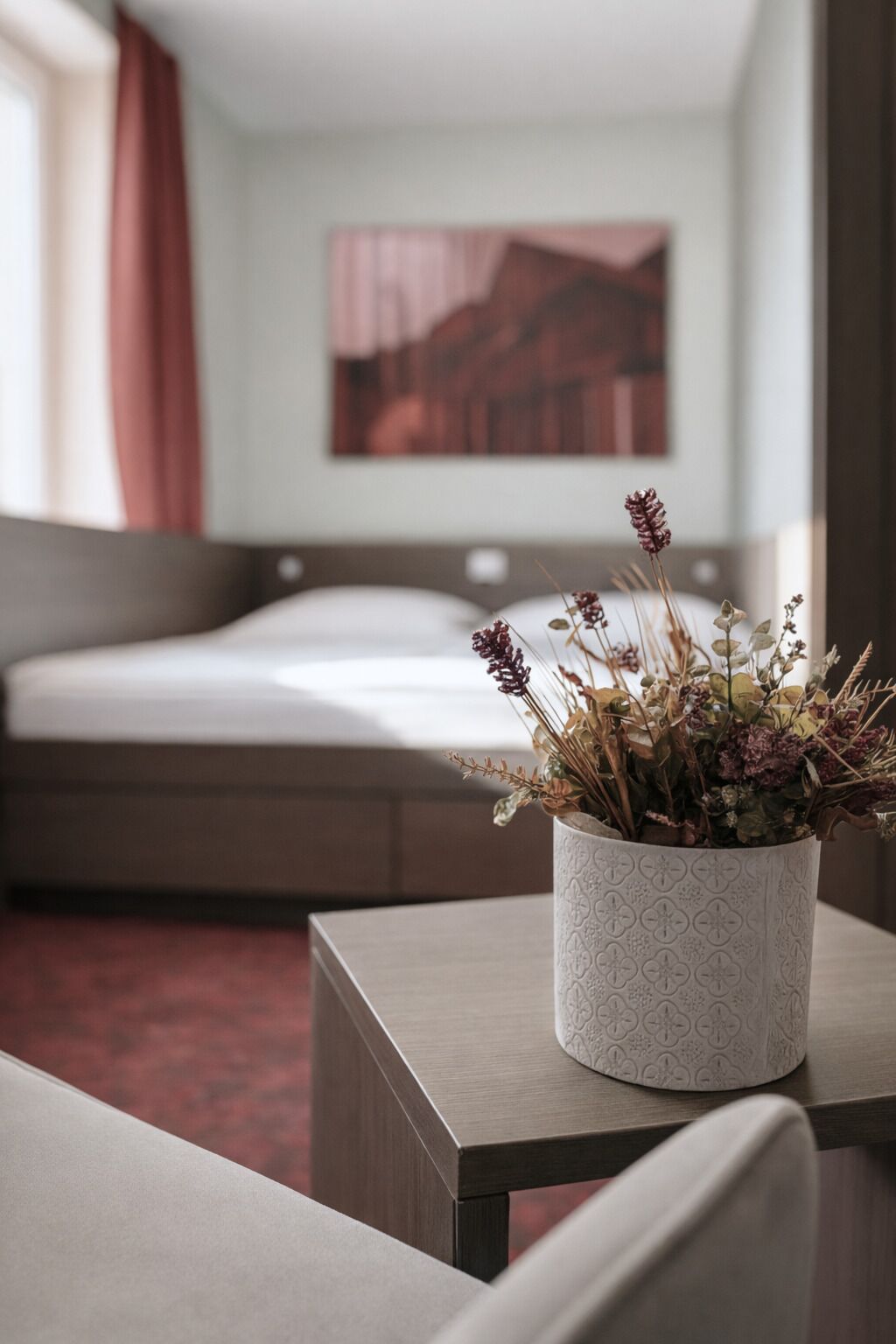 A hotel room with a bed, a bedside table with a potted plant, and a painting on the wall.