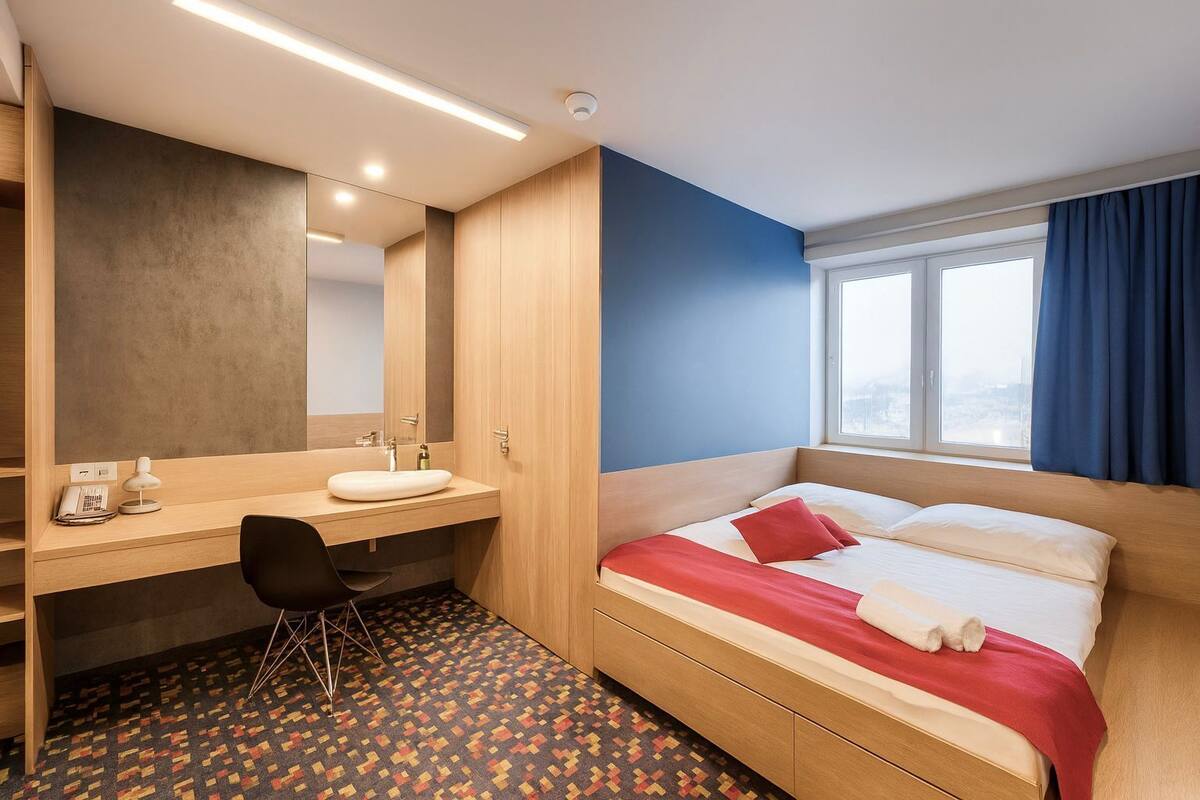 Hotellihuone, jossa on sänky, työpöytä tuolilla, peili, ikkuna verhoilla ja kuviollinen matto.