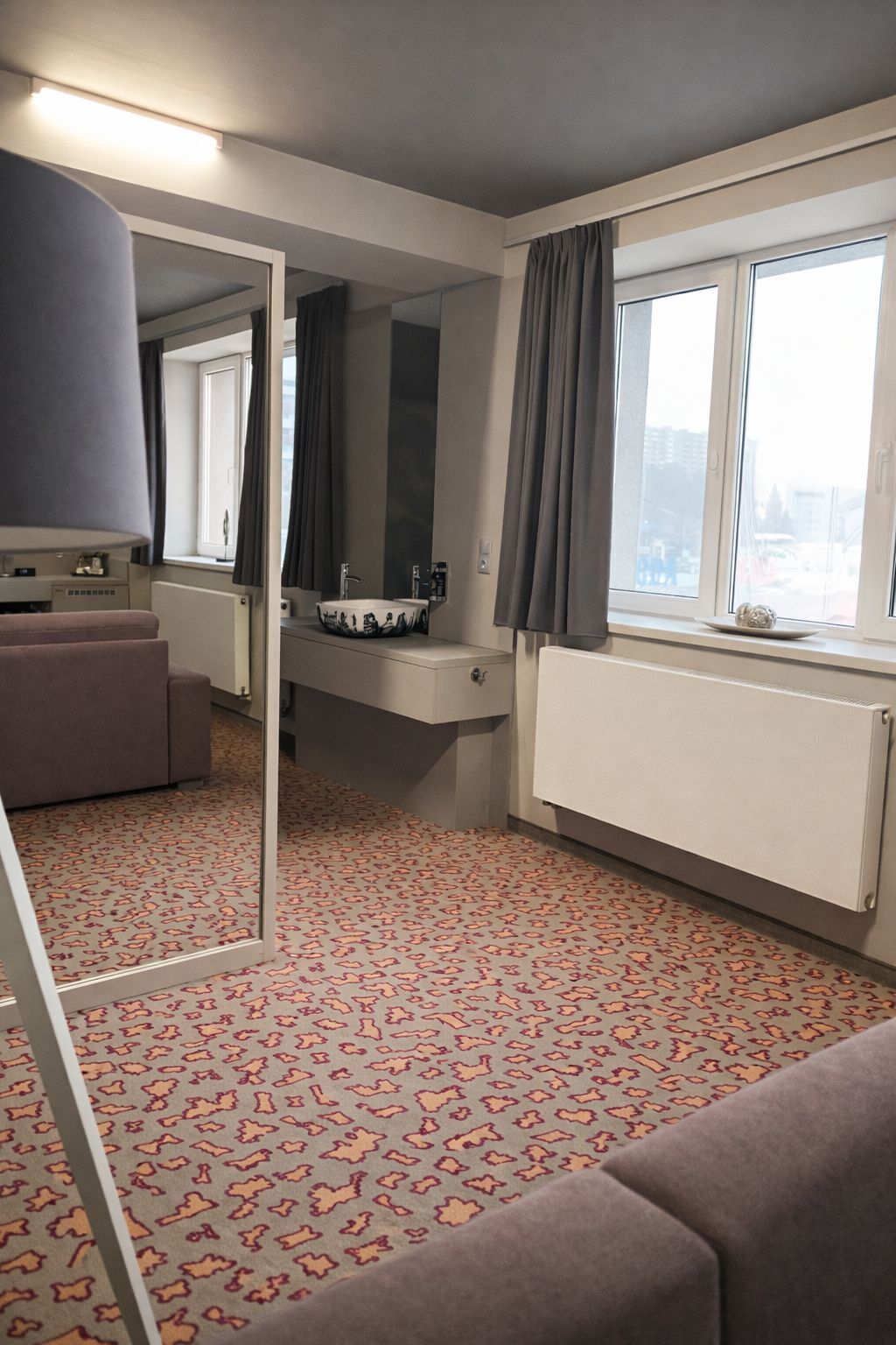 Una habitación de hotel con un espejo grande, un sofá, una ventana con cortinas y un radiador.