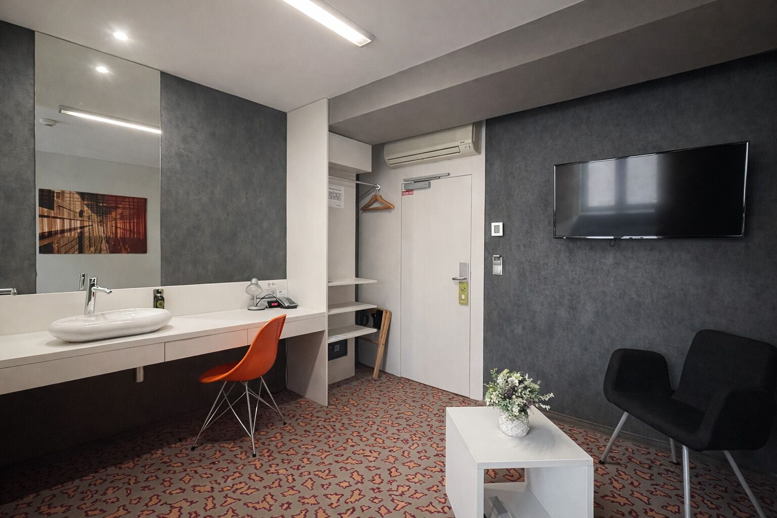 A modern hotel room with a large mirror, a white vanity with a sink, a flat-screen TV mounted on the wall, a small white side table with a vase, and a patterned carpet.