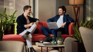 Two men sitting on a red sofa in a modern room with a lamp, a glass table, and potted plants.
