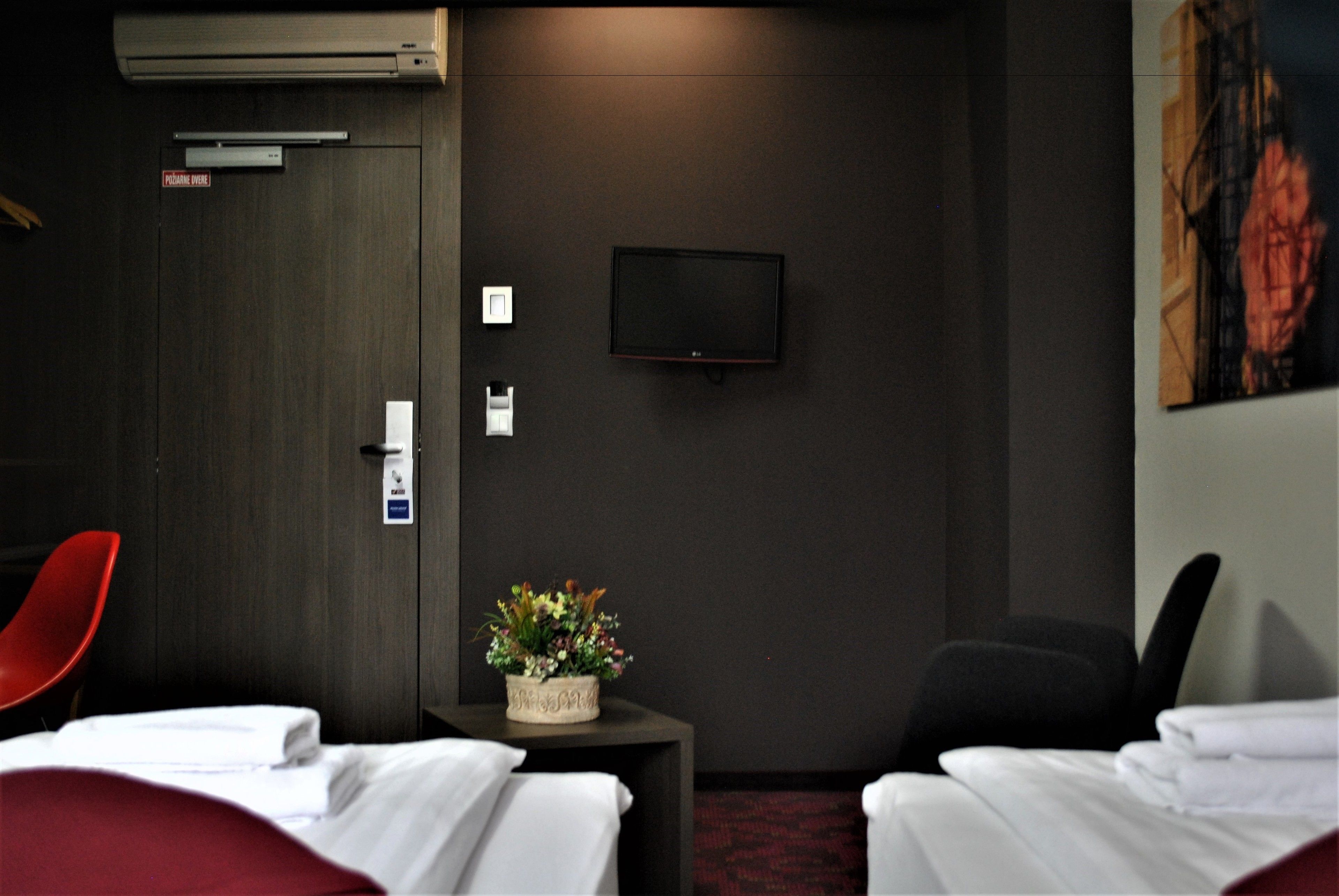 Une chambre d’hôtel avec deux lits, un téléviseur fixé au mur, un fauteuil rouge, une petite table avec un vase à fleurs et un tableau accroché au mur.