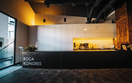 A reception area with a modern design, featuring a reception desk labeled 'ROCA KONGRES,' a potted plant, and a visible kitchen area in the background.