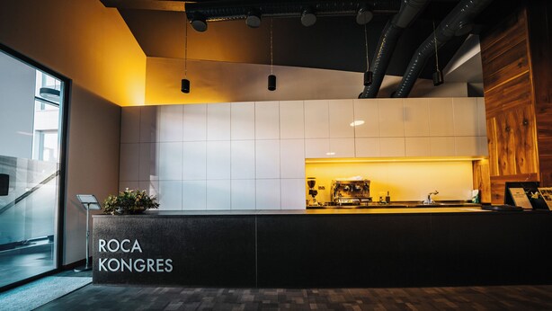 A reception area with a modern design, featuring a reception desk labeled 'ROCA KONGRES,' a potted plant, and a visible kitchen area in the background.
