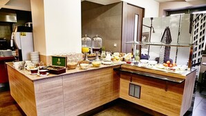 A breakfast buffet with various food items and beverages, a coffee station, and a view of a cityscape through a large window.