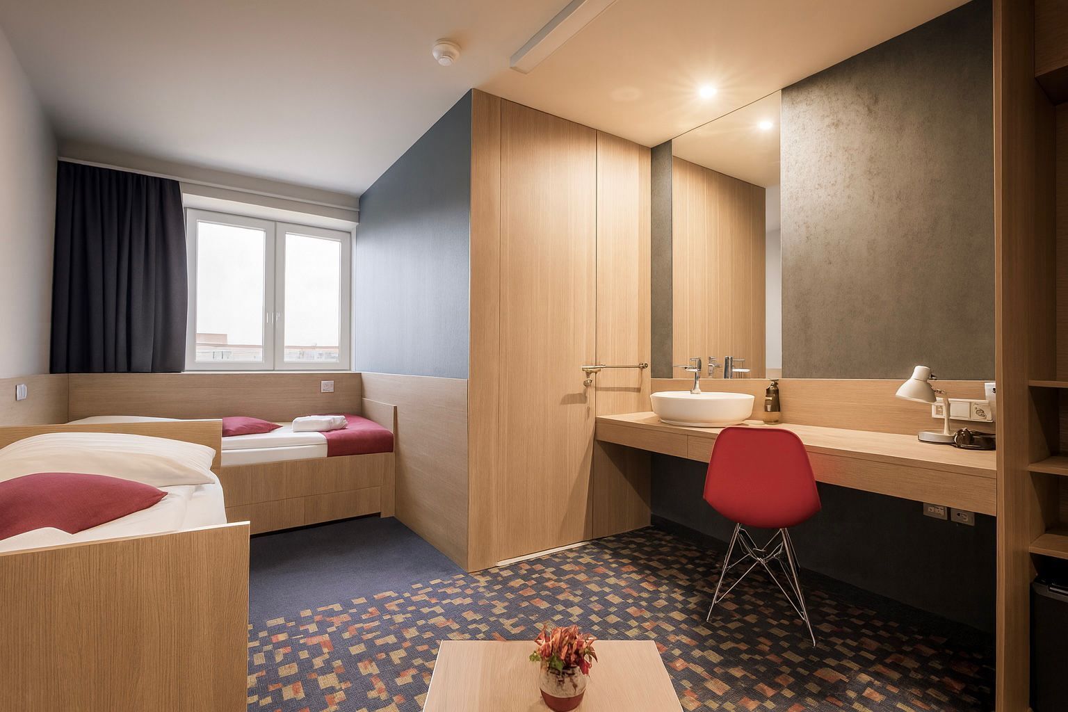 Une chambre d’hôtel de taille réduite, comprenant un lit, un bureau, une chaise, un lavabo et un miroir.