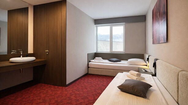 A modern hotel room with a large mirror, a wooden wardrobe, a bed with pillows, a bedside table, and a window with a view.