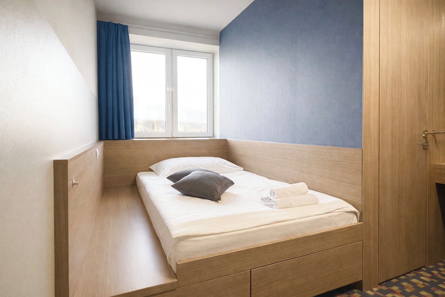 Una habitación pequeña con una cama individual, cabecera de madera, ventana con cortinas azules y una puerta a la derecha.