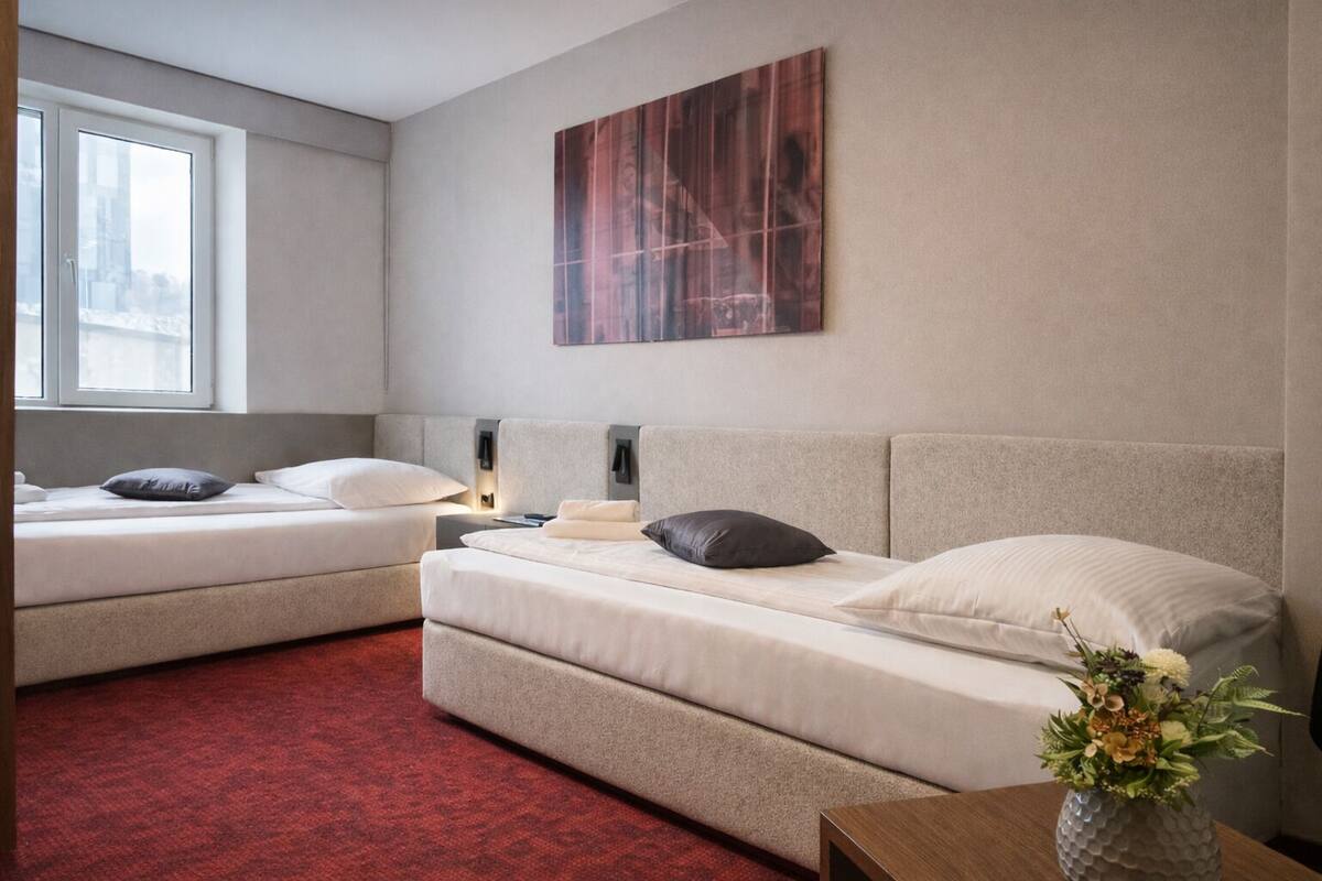 Hotellihuone, jossa on kaksi sänkyä, punainen matto, seinälle ripustettu taideteos ja maljakko kukkineen pöydällä.