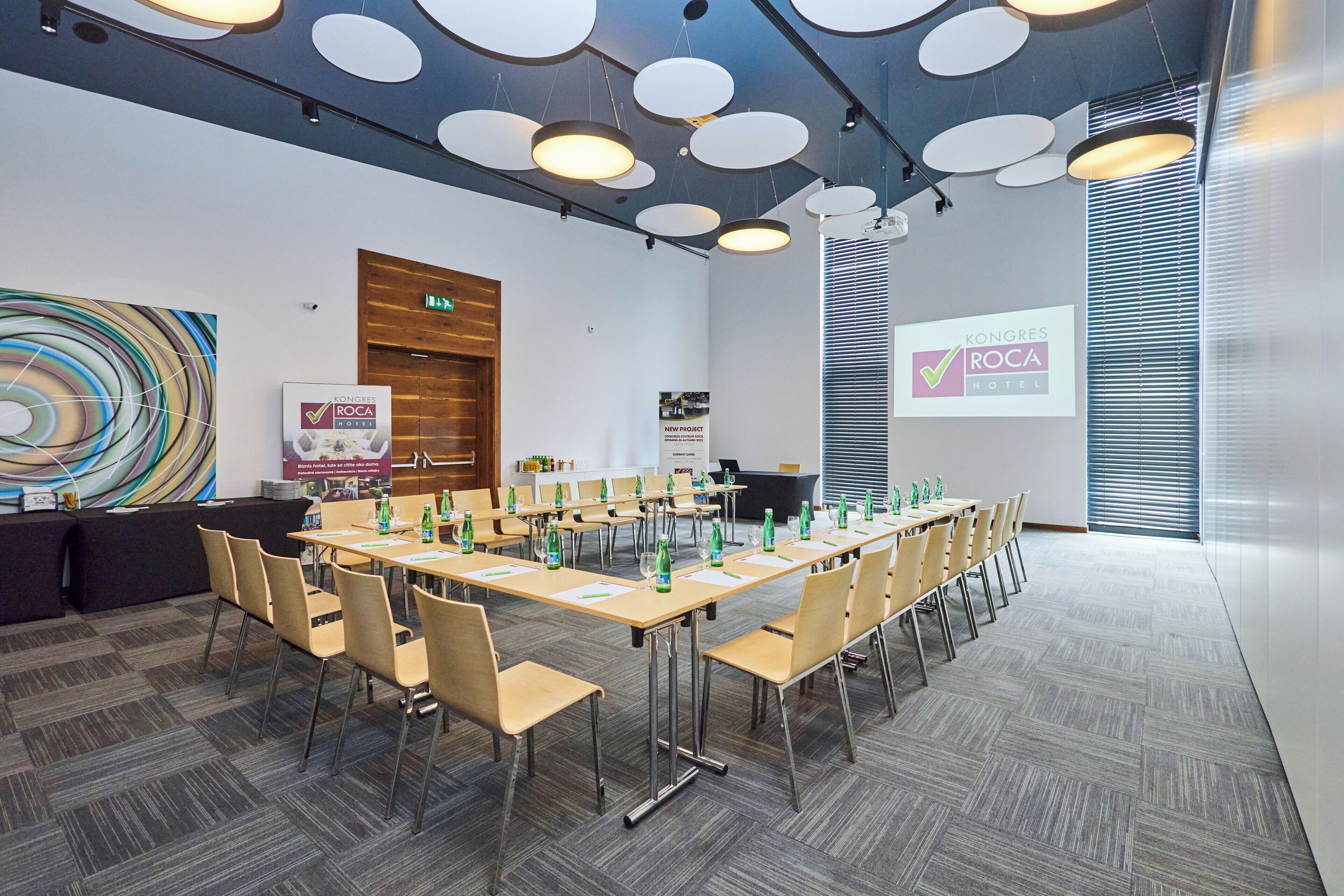 A conference room with a long table, chairs, and a projector screen displaying 'Kongres Roca Hotel'.