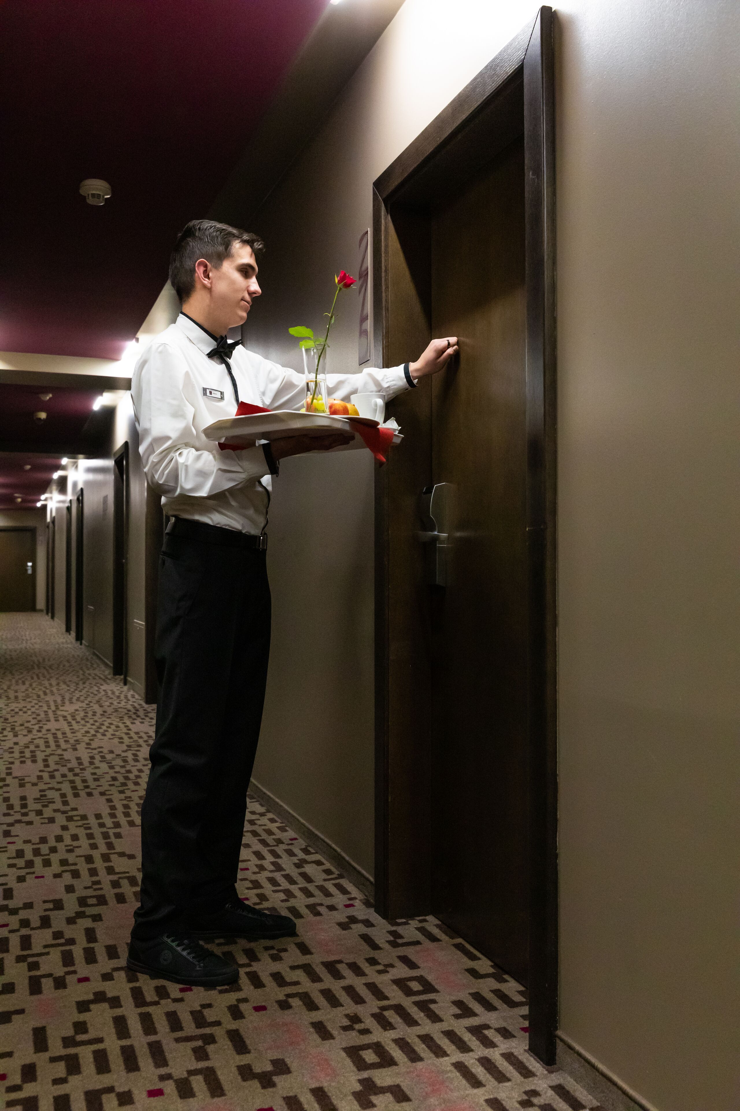 Un corridoio d'hotel con una persona che porta un vassoio con una rosa e un bicchiere di succo d'arancia.