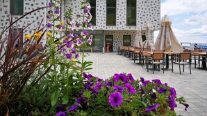 An outdoor seating area with tables and umbrellas, a building with a patterned facade, and purple flowers in the foreground. - Wellness Hotel Montis (Mozirje)
