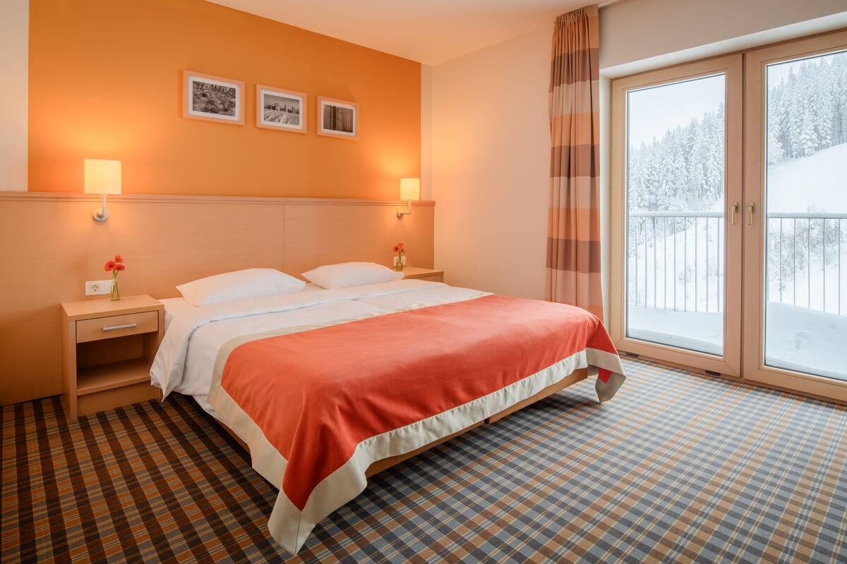 A hotel room with a bed, two bedside tables, a nightstand, a window with a view of a snowy landscape, and a checkered floor.