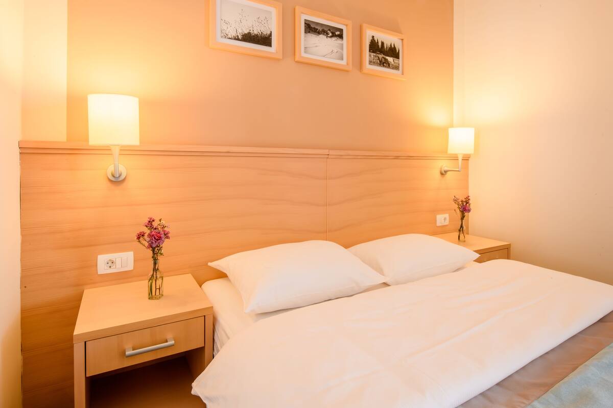A hotel room with a wooden headboard, two bedside lamps, a bed with white linens, a nightstand with a vase of flowers, and three framed pictures on the wall.