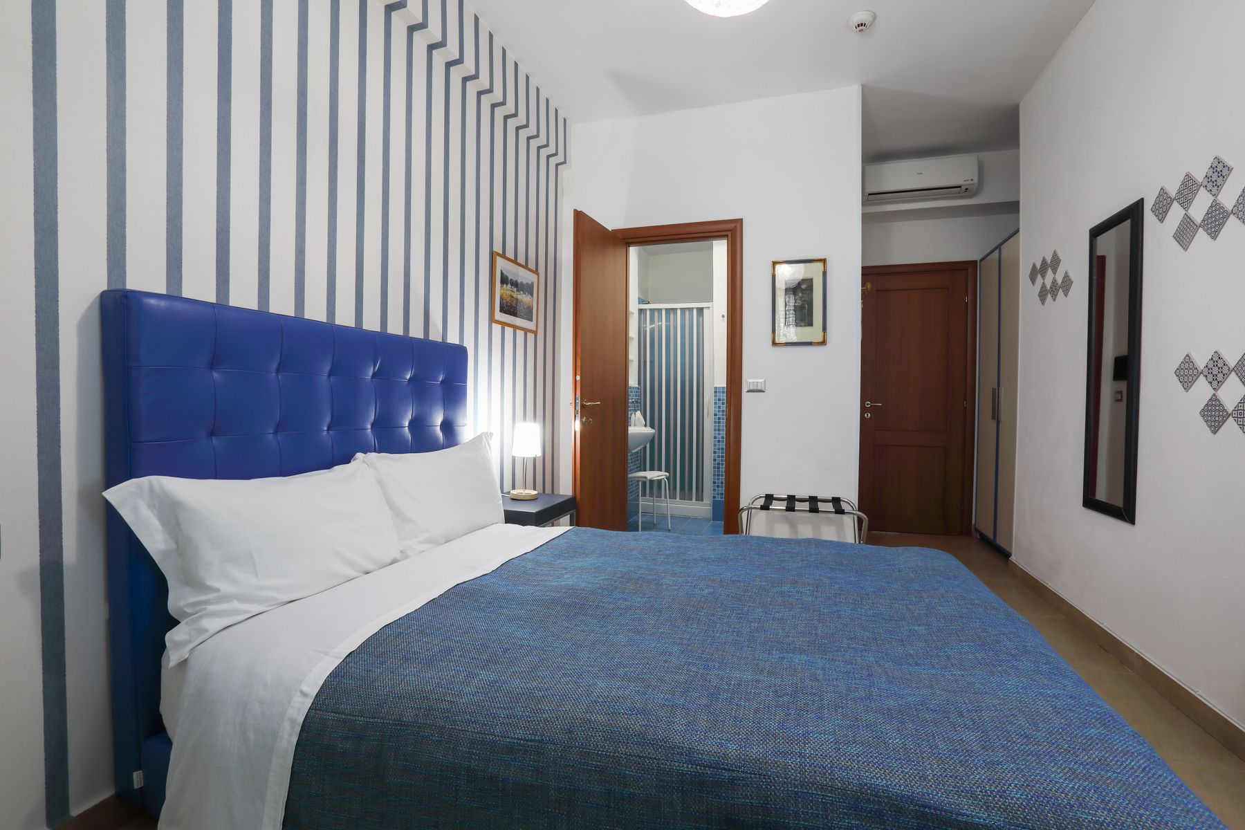 Une chambre à coucher avec une tête de lit bleue, un lit avec une literie blanche et bleue, une salle de bain avec une douche et un lavabo, et une armoire.