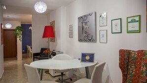 Una habitación de hotel moderna con una mesa redonda, dos sillas blancas, una lámpara roja y un sofá colorido. Hay diplomas enmarcados en la pared y una planta en la esquina.